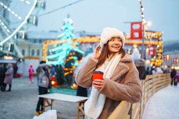 Beautiful woman  drinking hot coffee while walking in the  city market decorated with holiday lights. Holidays, rest, travel concept.
