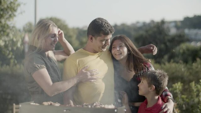 Happy man embracing and kissing his children at barbecue grill, his wife hugging him. Portrait of family looking at camera and smiling outdoors. Barbecue party and happy family concept