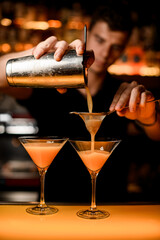 hand of bartender holds shaker with strainer and pours cocktail into martini glass.