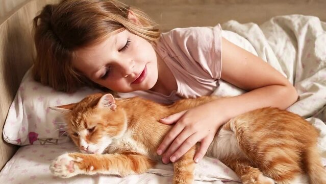 the child lies on the bed and strokes a beautiful red cat. child playing on bed at home