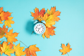Naklejka na ściany i meble Alarm clock on blue background and autumn colorful leaves. Daylight saving time end. Fall leaves. Autumn background. Back to school concept. Flat lay.