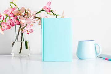 Blue book mockup, notepad, pen, cup, bouquet flower, pink orchids in glass vase. Home Office. Front view. Place for text, copy space, mockup