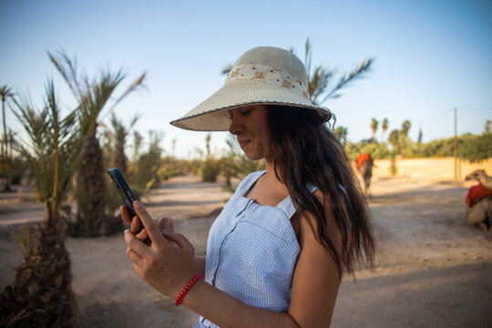 Happy sexy tourist with his hat using his cell phone in the palm grove of Marrakech, this desert place full of camels and dromedaries is very visited by tourists in Morocco.