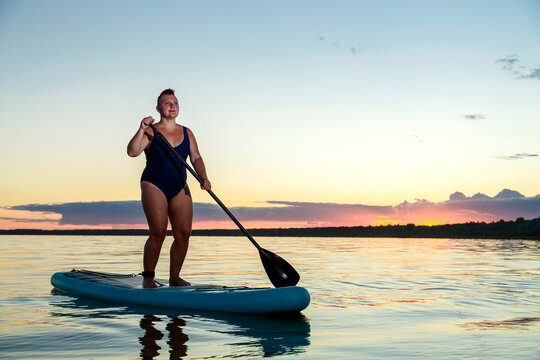a woman in a swimsuit with a mohawk on a sup board at sunset in the water.