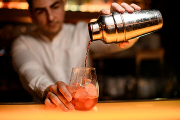 hand of male bartender gently holds shaker and pours cocktail into glass
