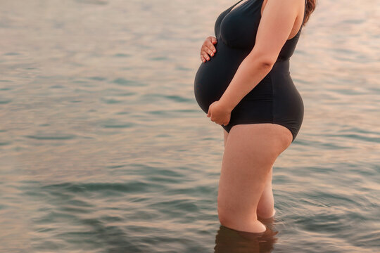 Motherhood and pregnancy. Young caucasian pregnant woman in swimsuit is standing in water and holding her tummy. Concept of water births and maternity