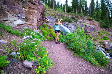Waterfall river on trail to Ice lake by Silverton, Colorado with hike path wildflowers and young woman taking photo picture of nature summer landscape