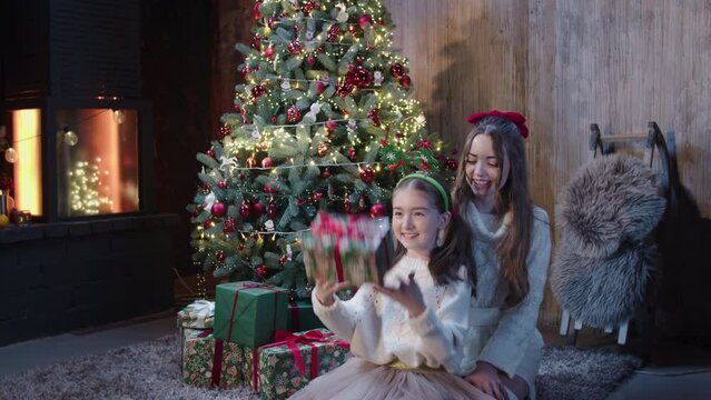 Image of a house magically decorated for Christmas. Two sisters dressed in white, next to a big Christmas tree full of lights and toys, preparing gifts for parents