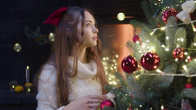Close-up image of a beautiful little girl with blue eyes, red bow in her hair, decorating the tree with red toys and looking at the tree. The charming Christmas atmosphere, with lights and candles