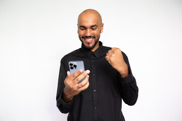Portrait of happy young man with phone making winning gesture over white background. Bearded businessman wearing black shirt reading newsletter on smartphone. Good news and mobile technology concept