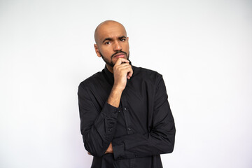 Portrait of thoughtful young man standing with hand on chin. Bearded businessman wearing black shirt looking at camera in doubt against white background. Uncertainty and planning concept