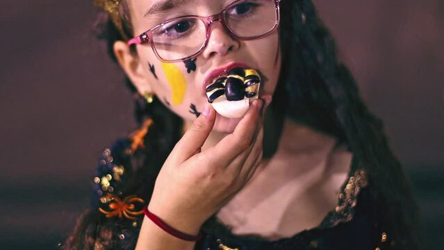 Beautiful caucasian girl with halloween makeup on face and princess dress eating egg with black olives spider celebrating halloween night outdoors, side view close-up in slow motion.