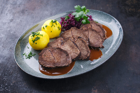 Traditionell geschmorter Sauerbraten vom Rind mit Blaukraut und Kartoffelklößen in würziger Dunkelbiersoße serviert als close-up auf einem Nordic design Teller