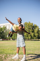 Portrait of African American man with little boy on his hands. Happy kid stretching hands flying and father standing on grassy field holding him on his hands. Parents love and active rest concept