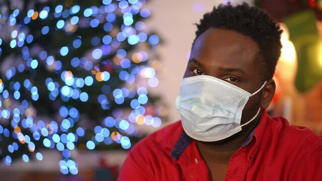 Close-up portrait of young African American man in coronavirus face mask looking back at Christmas lights and looking at camera shaking head no gesture. Guy posing at home on Covid-19 New Year