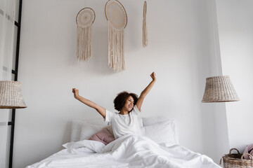 African girl in pajamas stertching in cozy bed and raising hands up at home. African american woman...