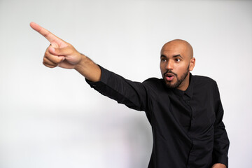 Portrait of amazed young man pointing finger at ad. Shocked multiethnic male model with bald head and beard in black shirt looking away, showing ads with open mouth. Shock, advertisement concept