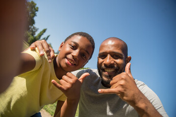 Portrait of happy African American man and boy having fun for camera. Father hugging his son, both looking at camera showing calling gesture with their fingers. Leisure and new technologies concept