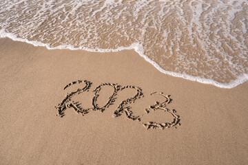 Hand-drawn numbers 2023 of new year on wet yellow sand on beach, copy space.