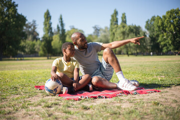 Naklejka na ściany i meble Portrait of African American man and boy resting on ground. Happy daddy and his son sitting on blanket, dad showing other people with his finger both looking there. Summer activity and leisure concept