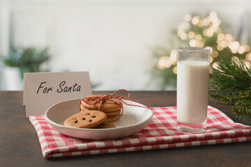 Christmas cookies in white plate and milk for Santa Claus in glass near Xmas tree. Festive tradition for winter holiday.