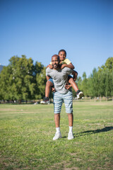 Full length portrait of happy father and his son posing together. African American man in casual clothes standing on field with boy on his back both looking at camera. Active rest, leisure concept
