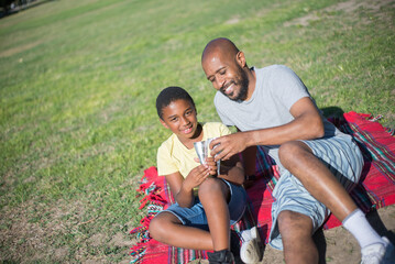 Portrait of young African American man and boy sitting on ground. Smiling happy father resting with boy in park both holding ice cream cones. Fatherhood, leisure and having rest together concept