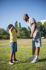 Side view of African American man and boy making faces on field. Happy dad and his cute son standing on grassy ground showing tongues to each other. Leisure and happy moments together concept