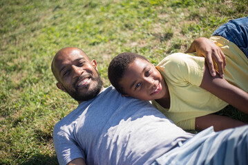 Top view of African American dad and son lying together on grass. Smiling man and boy hugging having rest after playing and looking at camera. Parents care, leisure and having rest together concept