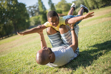 Portrait of happy dad and his son resting on field. African American bearded man lying on grass holding smiling boy with stretched hands on his knees. Leisure, sport activity and kids health concept