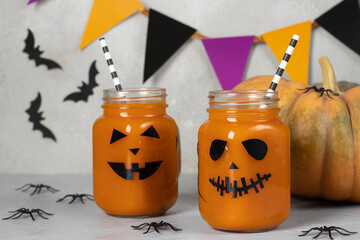 Pumpkin iced mocktails or smoothies in glass jars decorated with scary faces on light gray table....