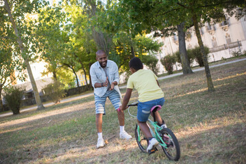 Fototapeta premium Portrait of happy African American dad and son riding bike in park.Smiling bald man pleased with kids progress clapping hands and looking at boy training to ride bike.Parenting and active rest concept