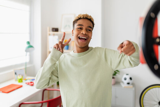 Cheerful young african american teenage boy recording video for social media. Millennial content creator male student having fun streaming online using mobile phone network app from domestic room