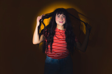 Haircare concept. Casually dressed caucasian teenage girl standing in studio in circle light, touching her healthy, long, dark, wavy hair and smiling. High quality photo