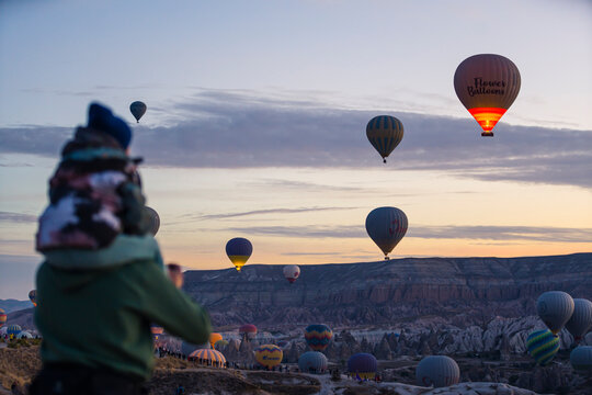 10.11.2022 Cappadocia, Turkey. Blurred parent holding his child piggy back in the foreground, watching stunning colourful balloons over Turkish town of Cappadocia. High quality photo