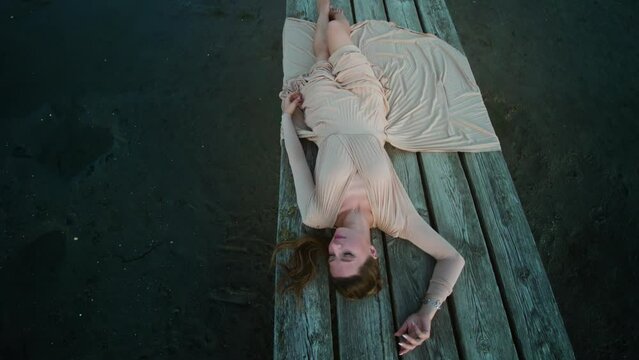 Young Fashion Model In Elegant Dress Lying On Wooden Pier On Banished Sand In The Evening