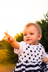 Toddler age 1 year summer sunny day outdoors. A little girl in a polka dot dress points her finger to the side and smiles.