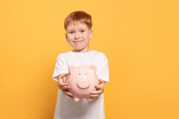 Fototapeta na wymiar Cute little boy holding ceramic piggy bank on orange background