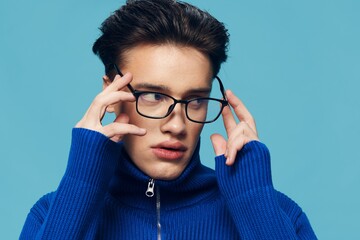 a close portrait of a young man in a blue jacket with black eyeglasses, standing on a light blue background holding them with his hand, looks away