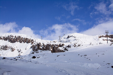 Tractor tracks crossing through prictine surface of the snow. Stunning winter landscape with snow blanketed mountain range and sky lift lines climbing up the slope. Mt Ruapehu, New Zealand