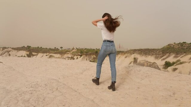 Slow motion, back view, attractive woman walking across rocky desert terrain and fixing her long brown hair. Country style, white baseball cap blue jeans brown boots. Confident gait