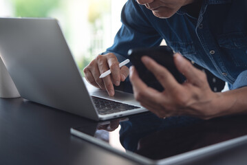 Close up of asian casual business man working on laptop computer, using mobile smart phone with digital tablet on table at home office