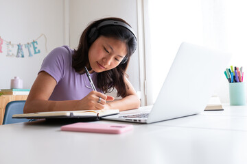 Asian female high school student writing on notebook. Chinese teen girl with headphone, studying at home using laptop.