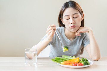 Obraz na płótnie Canvas Anorexia, unhappy beautiful asian young woman, girl on dieting, hand holding fork at broccoli in salad plate, dislike or tired with eat fresh vegetables. Nutrition of clean, healthy food good taste.