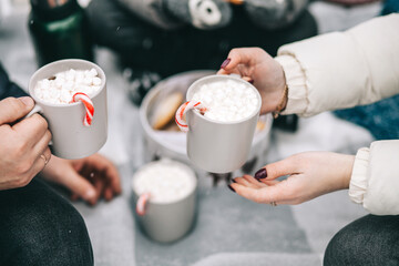 Mugs with cocoa and marshmallows in hands. Christmas picnic outdoors in the forest.