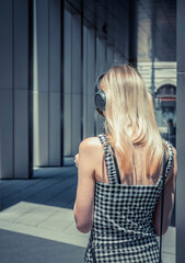 Fototapeta premium Rear view of a young blonde woman wearing over the ear headphones. Girl listening music on the street