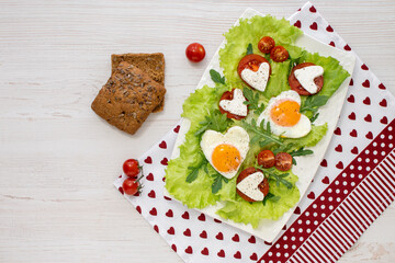 Diet festive breakfast on Valentine's Day. A plate of heart-shaped fried eggs, herbs, tomatoes and cheese on a white table covered with a napkin. View from above. Space for text.