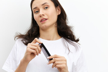 a sweet, pretty woman with long dark hair stands on a white background in a white T-shirt and holds a black comb in her hands, looking pleasantly at the camera