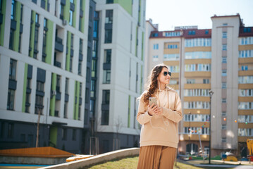 Young stylish smiling long-haired brunette uses a cellphone and headphones on the street. Woman talking on video chat. Urban city background.