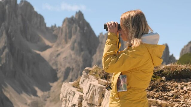 Female tourist looks through binoculars under blue sky. Woman admires rocky mountains of Italian Apls near Tre Cime di Lavaredo closeup backside view